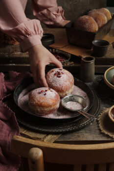 pączki ułożone na rustykalnej tacy lub talerzu posypane cukrem pudrem, w tle osoba w różowej bluzce, która sięga po jednego pączka, fotograf kulinarny lublin pyszny kadr kinga ciszewska