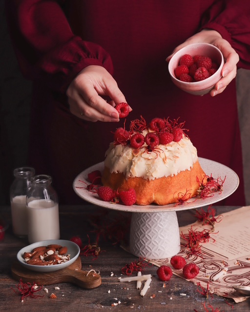 babka wielkanocna stoi na paterze, jest ozdobiona lukrem i świeżymi malinami, w tle stoi osoba, która dekoruje ciasto, fotografia kulinarna pyszny kadr kinga ciszewska