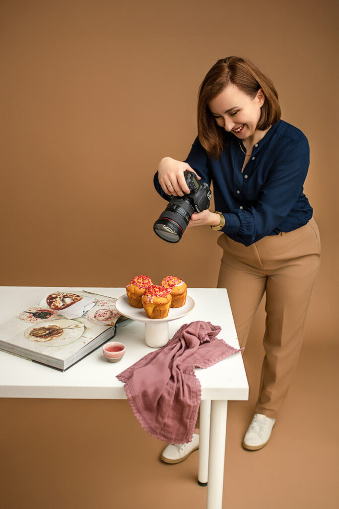 Fotograf kulinarny Kinga Ciszewska podczas pracy – uśmiechnięta, fotografuje słodkie wypieki na białym stole. W tle książka kucharska i stylowe rekwizyty. Profesjonalna sesja kulinarna w ciepłych, naturalnych kolorach.