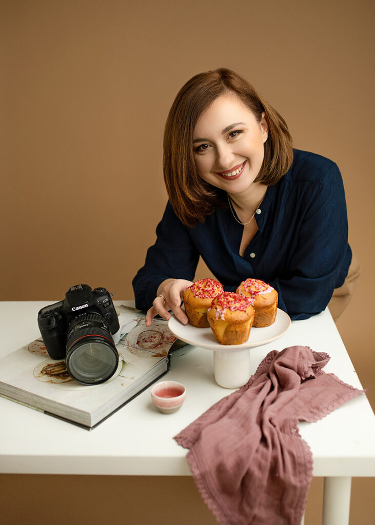 Fotograf kulinarny Kinga Ciszewska uśmiecha się do obiektywu, trzymając talerz z apetycznymi wypiekami. Na stole znajduje się aparat Canon, książka kucharska i stylowe rekwizyty. Profesjonalna fotografia kulinarna w ciepłych, naturalnych barwach