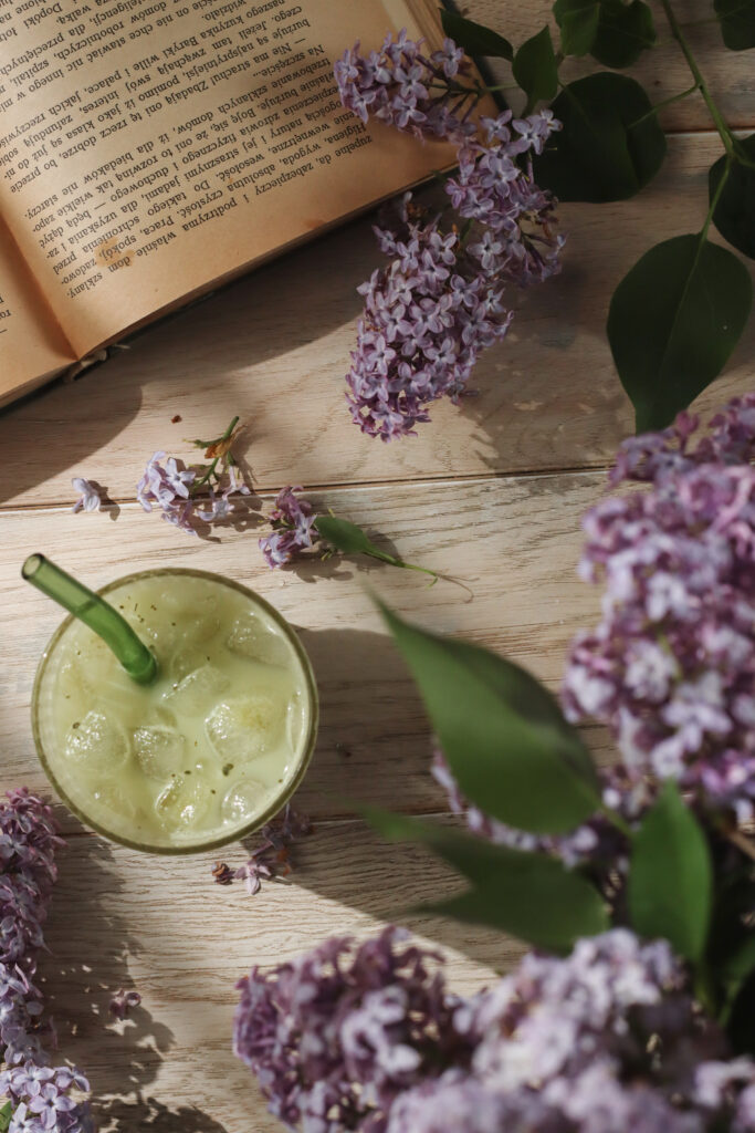 Szklanka zielonej matcha latte z lodem i zieloną słomką, ustawiona na jasnym drewnianym stole w towarzystwie bzu i otwartej książki - fotografia jedzenia w stylu wiosennym, z naturalnym światłem.