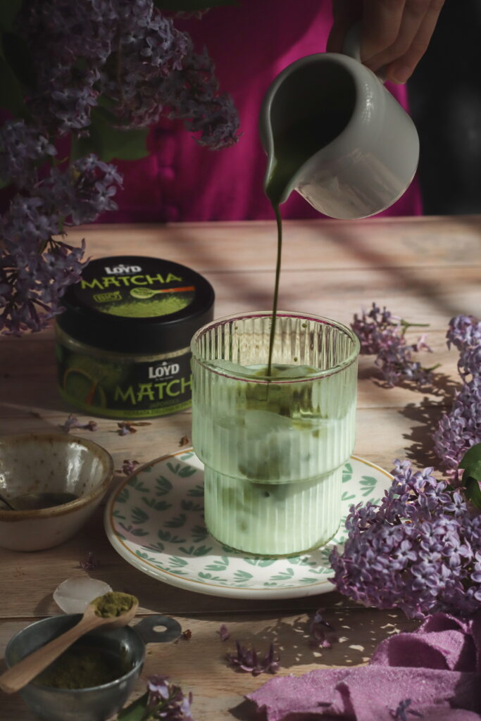 Nalewanie matchy do szklanki z mlekiem i lodem, otoczonej bzem i stylizacyjnymi dodatkami; w tle opakowanie herbaty matcha LOYD - fotografia jedzenia z naturalnym światłem w stylu wiosennym.