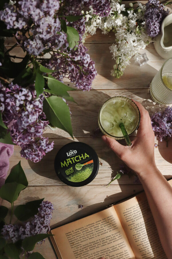 Szklanka matcha latte z lodem trzymana w dłoni na jasnym drewnianym stole, otoczona kwitnącym bzem, książką i opakowaniem herbaty matcha LOYD - stylizowana fotografia jedzenia z naturalnym światłem.